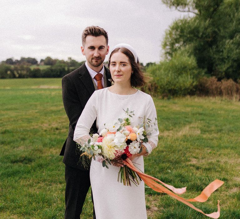 Groom in three piece suit with light coloured shoes embraces the bride from behind with the bride in a high neck three quarter length sleeve wedding dress with headbands and homegrown DIY wedding bouquet 