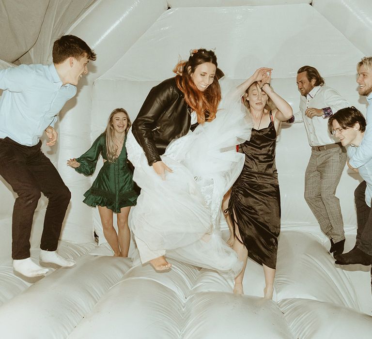 Bride in personalised leather jacket jumping on the bouncy castle with some wedding guests during the wedding reception 