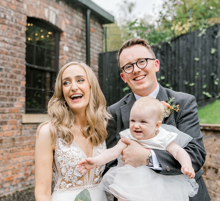 Bride in Mikaella Bridal wedding dress stands beside her groom and their daughter in white dress with tulle skirt