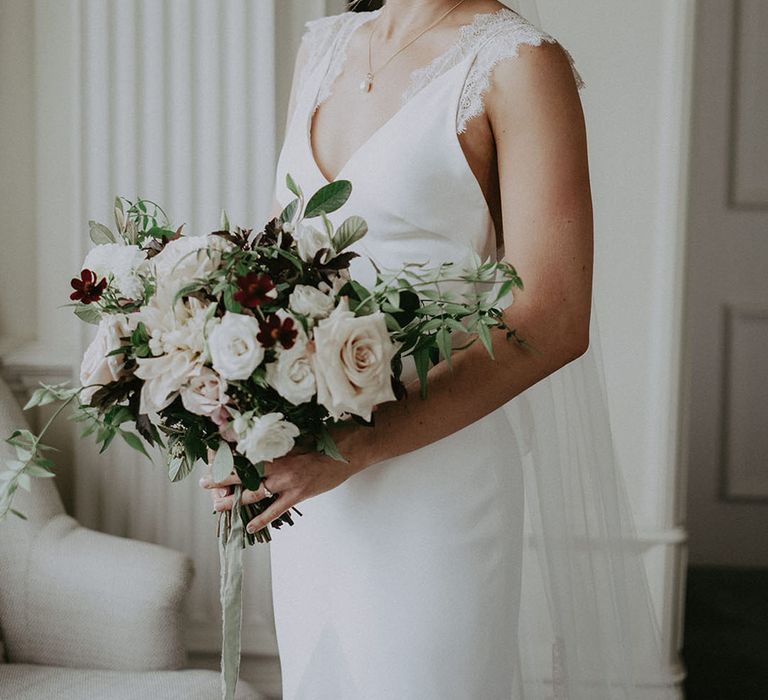 Bride holds classic bridal bouquet complete with white florals and green foliage tied with raw edge ribbon