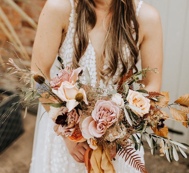 Gorgeous bride with a half up half down braided hairstyle with pearl stud earrings in a plunging Made With Love wedding dress holding an autumnal bouquet 