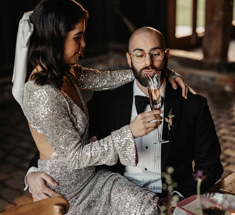 Bride in open back long sleeve silver sparkly wedding dress giving groom in classic black tuxedo and black bowtie a glass of champagne 