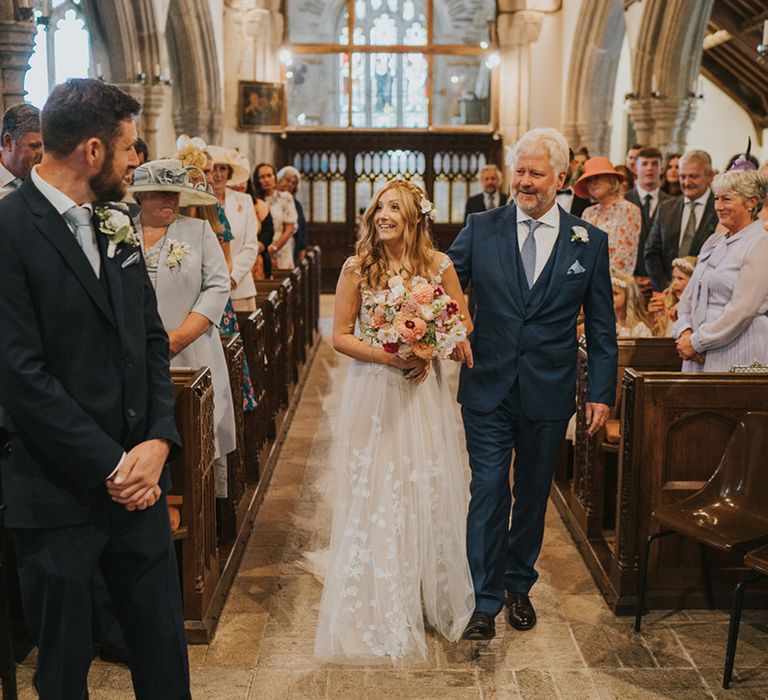 Father of the bride in blue suit with pale blue tie walks with the bride down the aisle to the groom in a navy suit standing at the altar 