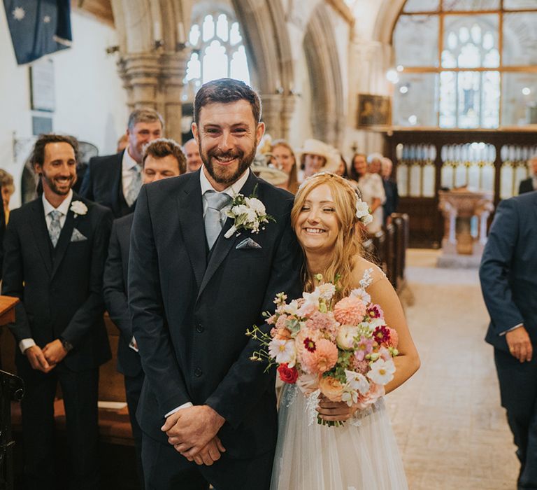 Groom in dark blue suit with light blue tie and white flower buttonhole smiling with the bride for church wedding in Devon