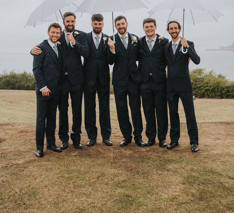 Groom and groomsmen in matching navy suits with light blue ties standing under umbrellas at rainy wedding 