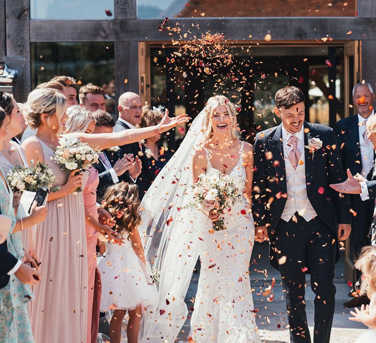 Groom in three piece suit with pink tie walking with the bride in a fitted lace wedding dress walking along as confetti is thrown over them 