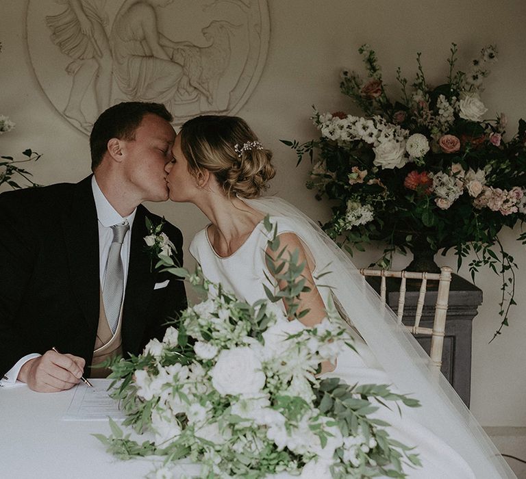 The bride and groom share a kiss as they sign the wedding register together with white and pink wedding flowers surrounding them 