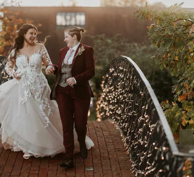 Bride in long sleeve lace illusion sleeve wedding dress with overlay walking over a bridge at Bassmead Manor Barns with bride in maroon bridal suit with grey waistcoat, dusky pink tie and autumnal dried flower boutonniere 