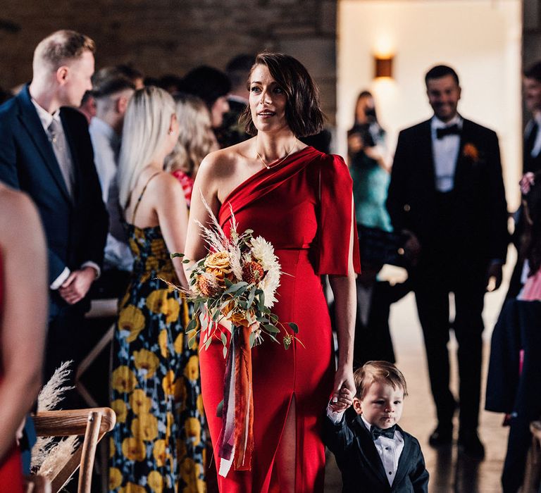 Bridesmaid in red satin bridesmaid dress walking down the aisle with page boy in mini black tuxedo holding an orange flower bouquet 