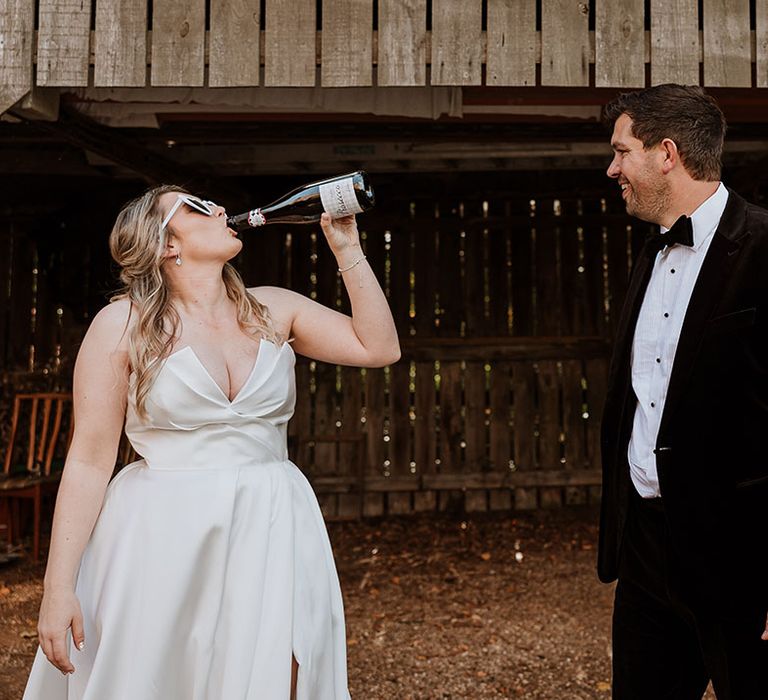 Bride in strapless Suzanne Neville wedding dress wearing white bridal sunglasses drinking champagne from the bottle with the groom in black velvet tuxedo 