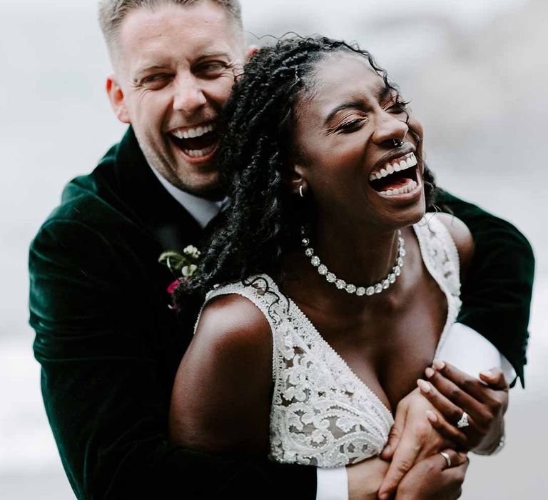 Bride in v neck sleeveless lace Pronovias wedding dress and silver sparkly bridal jewellery smiling with groom on Tunnels Beaches in bottle green velvet grooms blazer