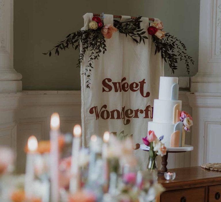 Spring themed wedding tablescape and three tiered pastel wedding cake beside large white wedding banner 