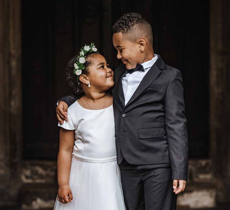 Flower girl wearing short sleeve satin dress and flower crown looking up at page boy in black suit and bowtie at Lains Barn wedding