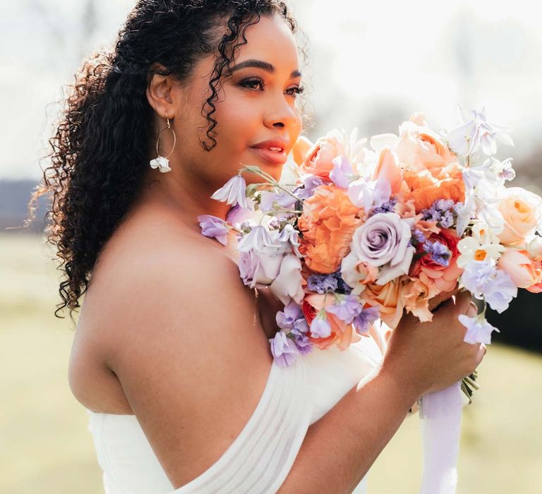 Bride in off the shoulder wedding dress and silver bridal crown holding orange, lilac and peach mixed flower bridal bouquet holding bridal bouquet with peach garden roses, white peonies, lilac sweet peas, bluebells and wildflowers tied with lilac ribbon