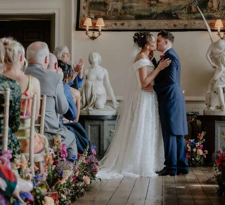 Groom in navy groom morning suit with grey ties and blush rose boutonniere kissing bride in Suzanne Neville off shoulder 3D floral applique wedding dress at the alter 