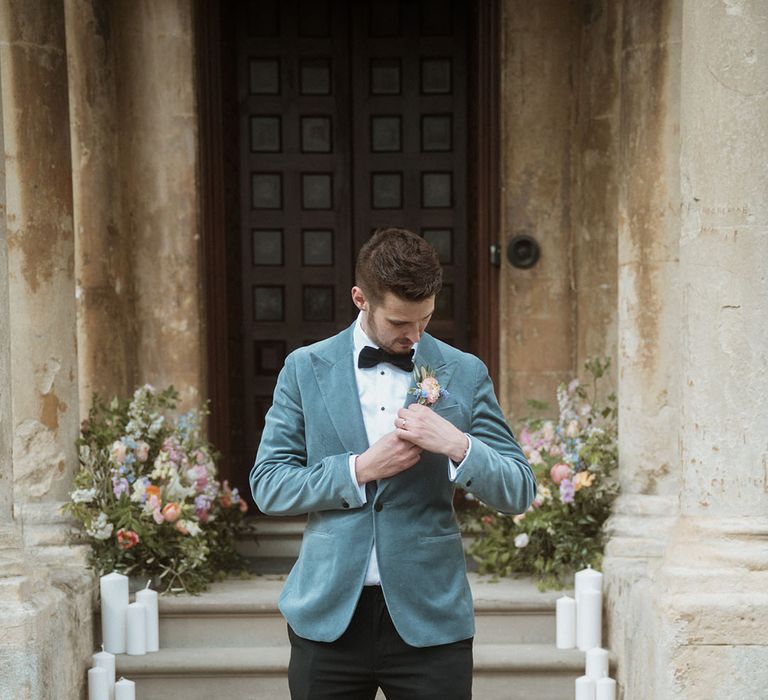 Groom in black trousers, black bow tie, blue velvet blazer and pastel floral buttonhole 