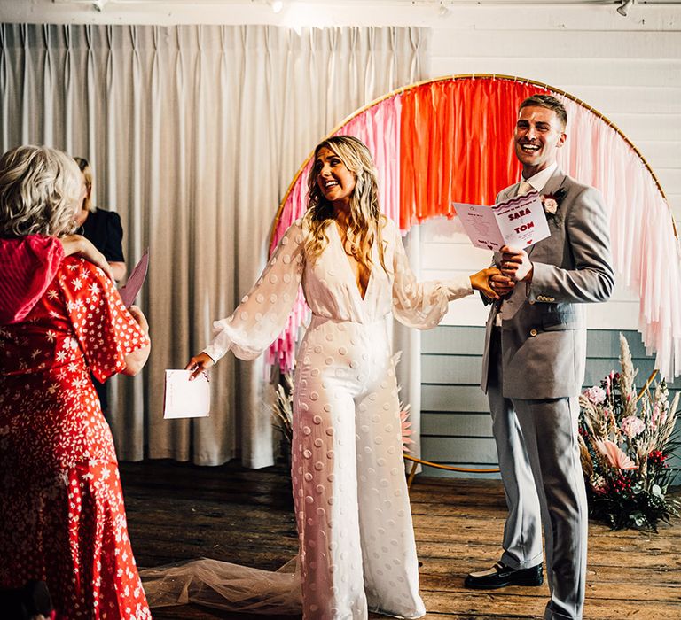 Bride in a jumpsuit and groom in a light grey suit at the streamer and hoop moongate altar at East Quay whitstable wedding venue 