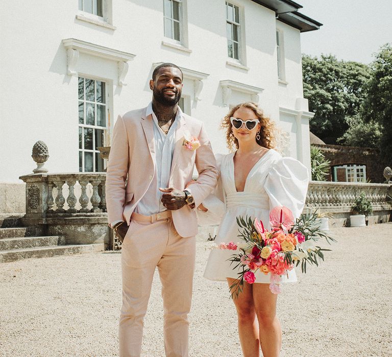 Bride in puff sleeve short wedding dress carrying white heart shaped retro wedding sunglasses with pink bouquet with groom in pink suit and buttonhole 