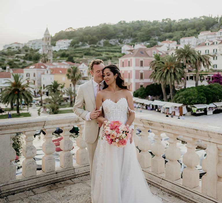 groom in beige linen suit embraces bride in 3D lace off-the-shoulder wedding dress from behind while bride holds pinka nd blush wedding bouquet with Old Town Hvar in the background