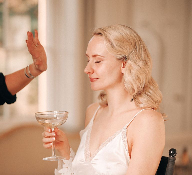 Bride in white satin pyjamas sits getting her makeup done for the wedding day 