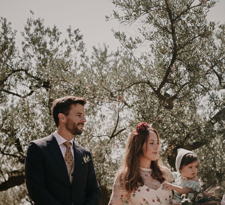 The Bride and Groom stand in an olive grove holding their child