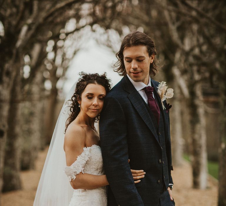 Portrait of the bride with brown curly hair in an off the shoulder lace wedding dress embracing her groom in a navy check suit 