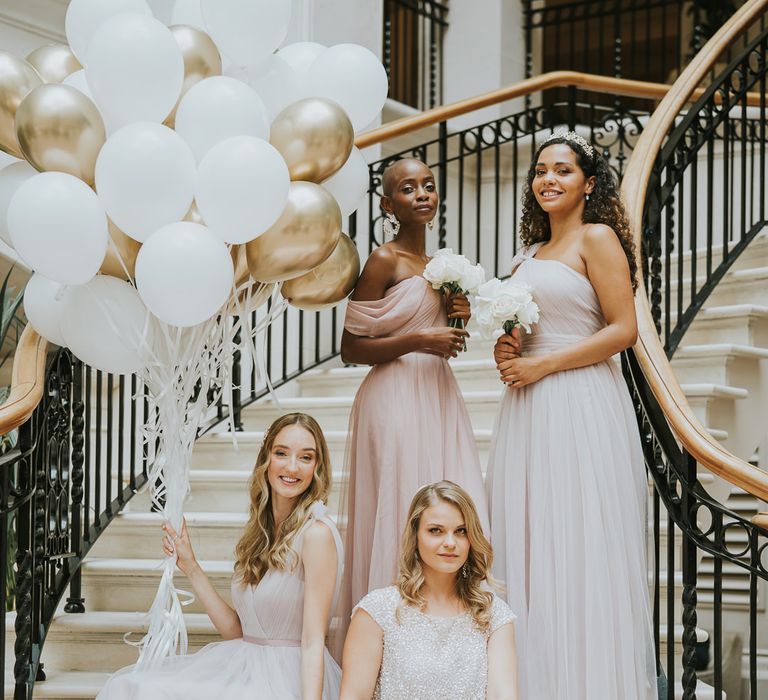 Bride in sparkly bridal separates sitting on the steps at The Landmark London with her bridesmaids in chiffon dresses holding metallic biodegradable balloons on strings 