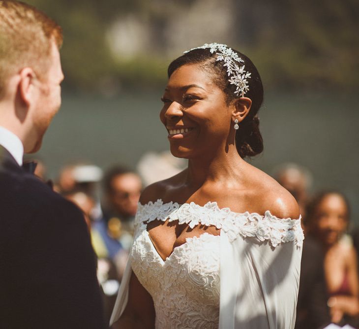 A bride with an off the shoulder dress smiles at the groom at the altar. She has her hair in a low bun and wears a sparkly hair accessory. 