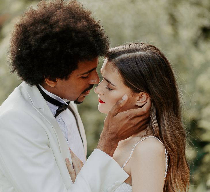 Black groom with afro hair and white tuxedo jacket holding his brides face with long brown hair and red lipstick