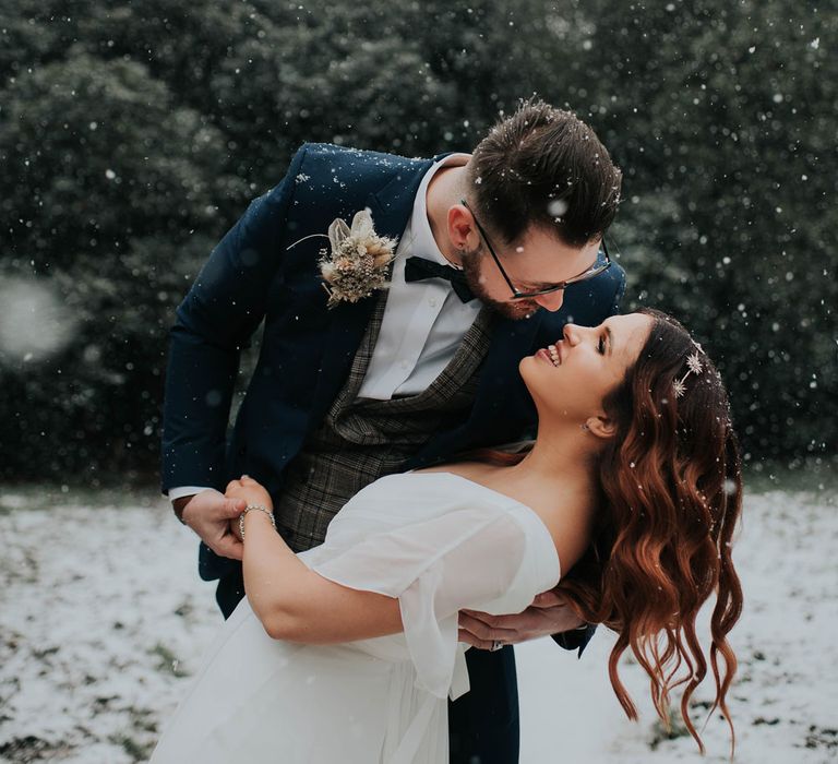 Groom in navy suit and bowtie dancing with bride with glam waves at snowy wedding 