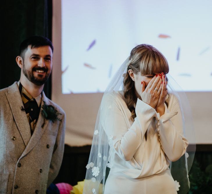 Stylish bride in a wrap jumpsuit with button detail on the sleeves covering her face with excitement 