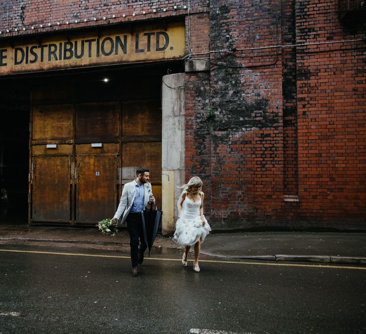 Bride and groom crossing street in an industrial area. Bride holds up dress and groom holds black umbrella in one hand and bride's bouquet in the other.