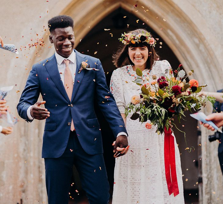 Confetti moment with the bride in a Broderie Anglaise wedding dress and flower crown and groom in a navy suit 