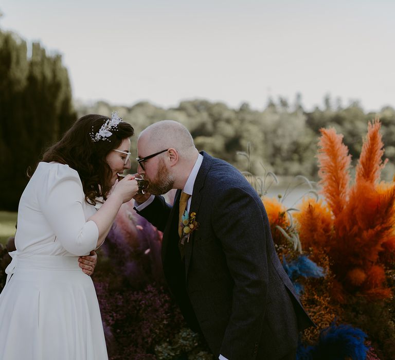 Bride in Rime Arodaky wedding dress and bridal headband drinks out of silver cup with groom in navy suit and yellow tie in humanist wedding ceremony