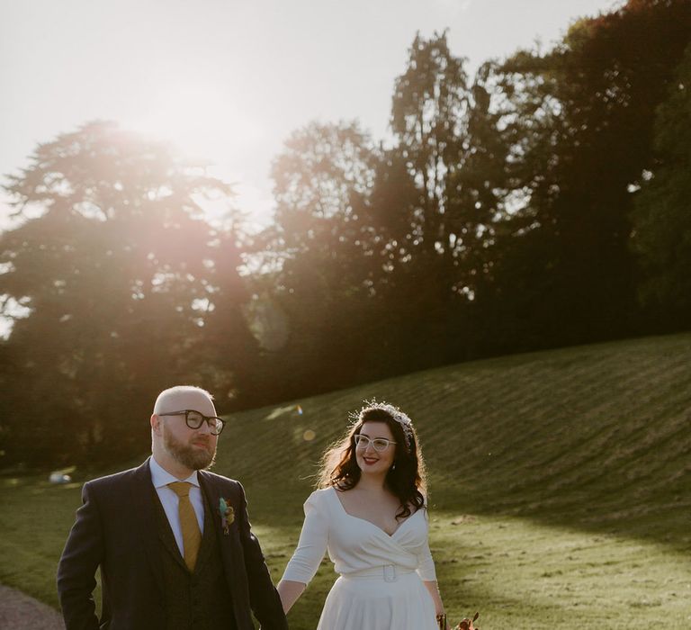 Groom in navy suit and yellow tie walks with bride in white cat eye glasses and bridal headband holding white and pink rose and pampas grass bouquet