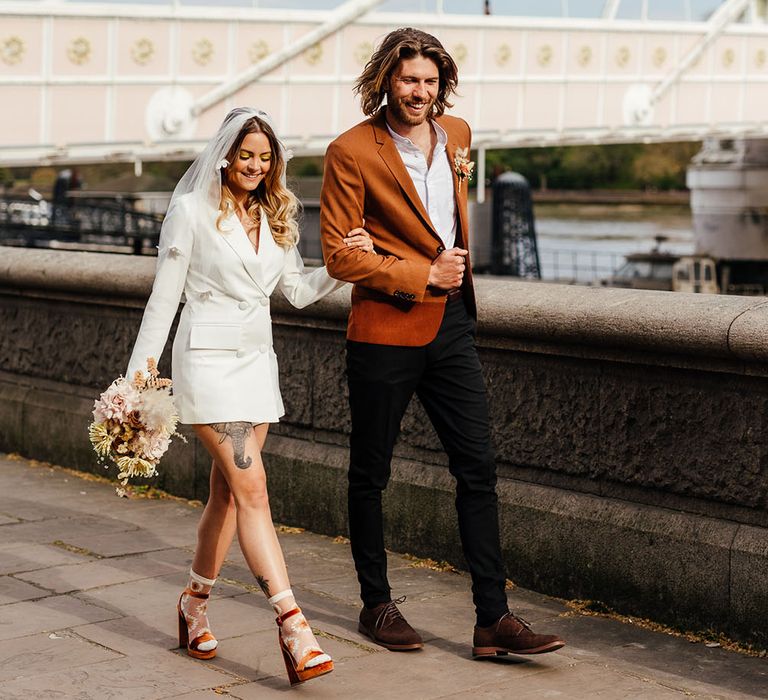 Bride in a white blazer dress and appliqué veil walking across Albert Bridge in Chelsea with her groom in a rust blazer 