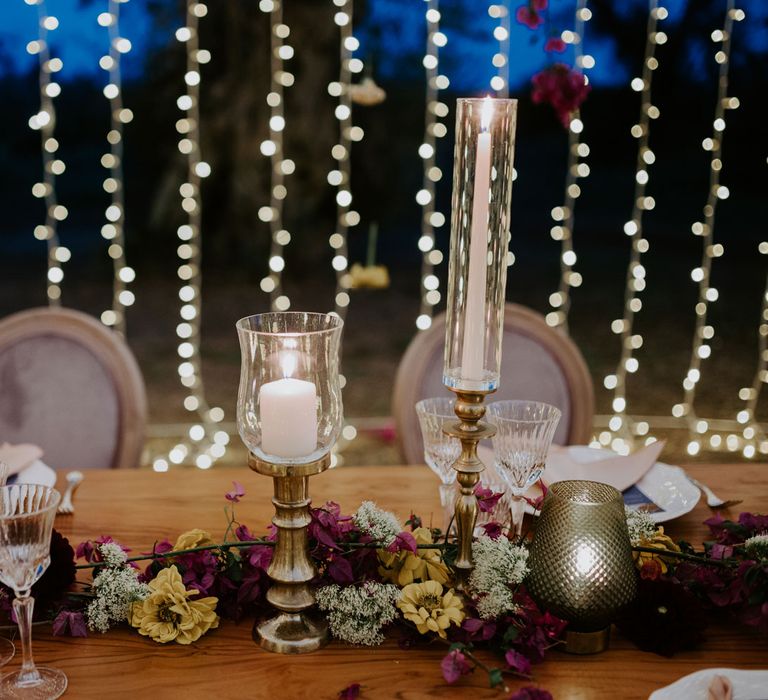 Glass and bronze candle holders, cut crystal glasses and multicoloured floral table garland on rustic wooden table with fairy light backdrop at enchanted forest wedding in Italy