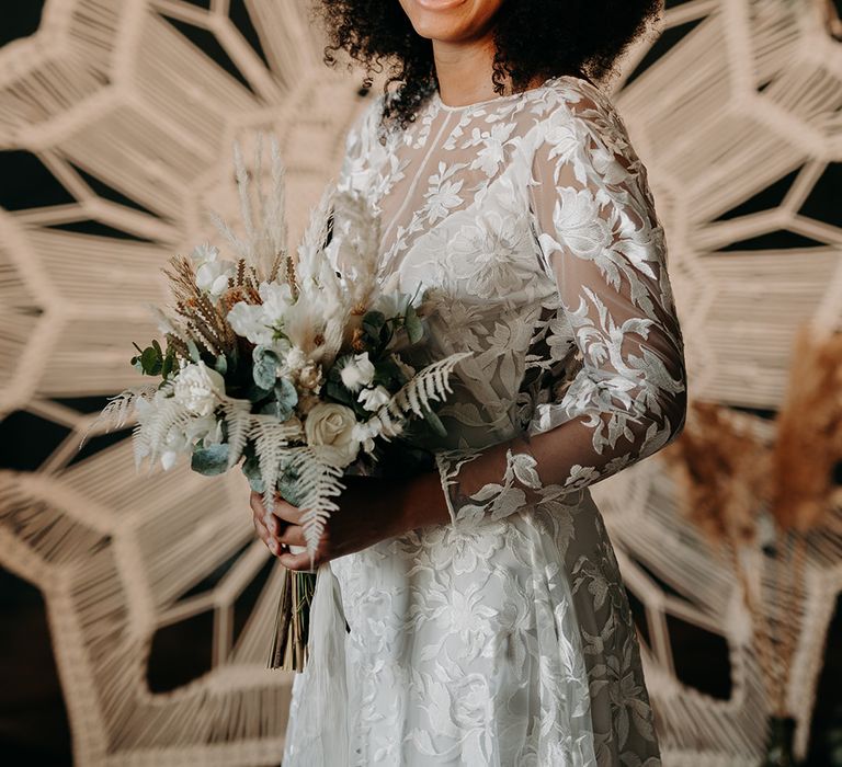 Black bride wears hair naturally and lace boho gown whilst holding floral bouquet