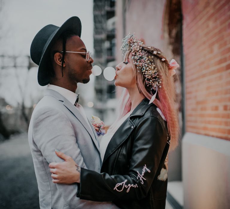 Groom in pale blue wedding suit and black hat blowing bubbles with his bride in a leather jacket 