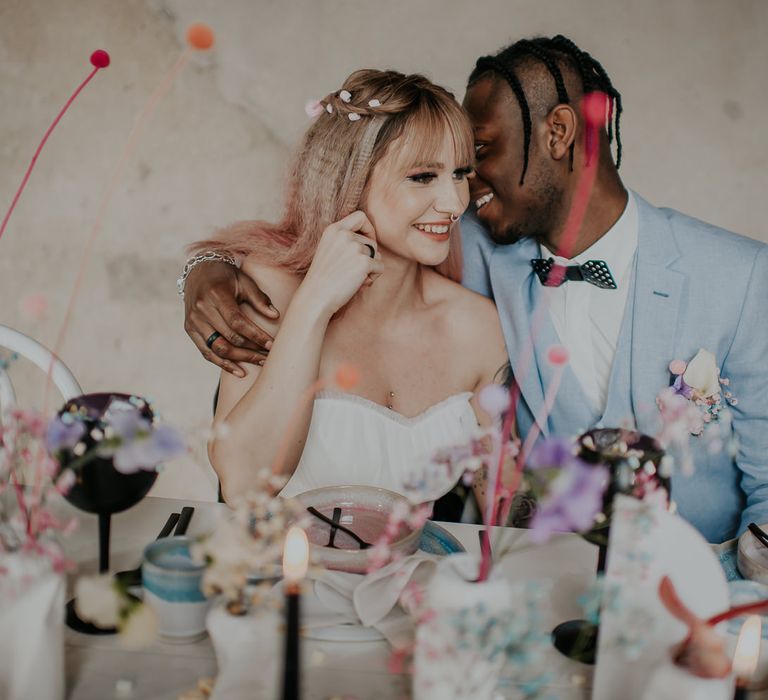 Groom in a pale blue wedding suit with black metal bow tie whispering sweet nothings into his brides ear with pink ombre hair 