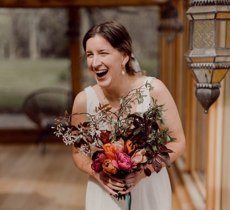 Laughing bride in V neck Charlie Brear wedding dress and chapel length daisy applique veil holds multicoloured bridal bouquet with foliage and green ribbon