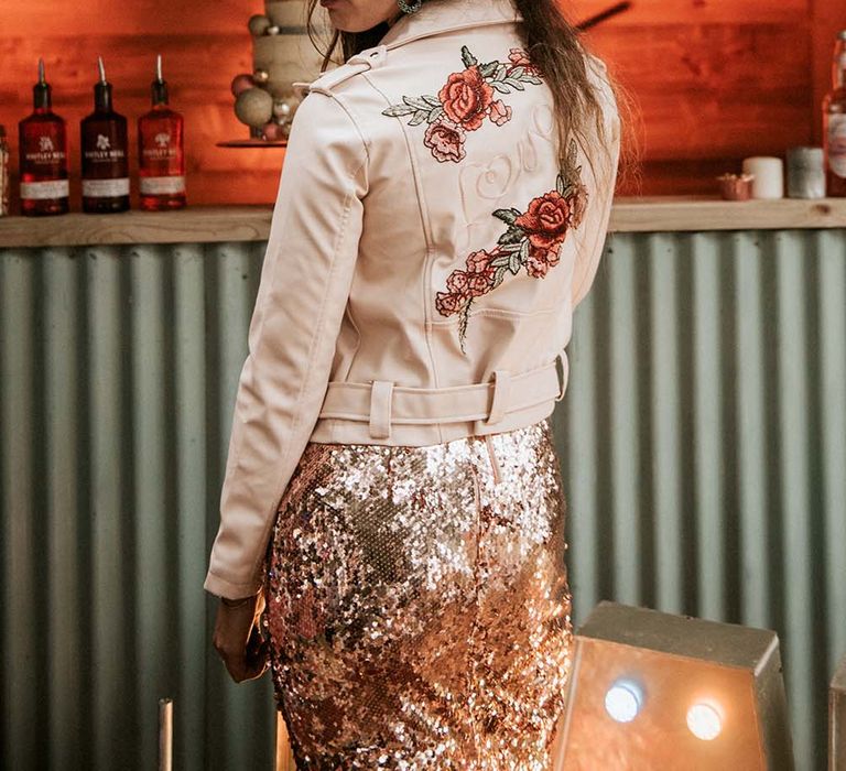 Bride in rose gold sequin wedding dress and face glitter wears blush pink leather jacket with embroidered rose detailing on the back for festival themed wedding