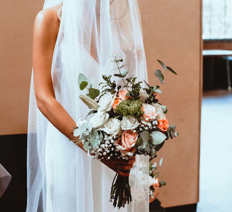 Bride in a cathedral length veil holding a peach and white rose bouquet with foliage 