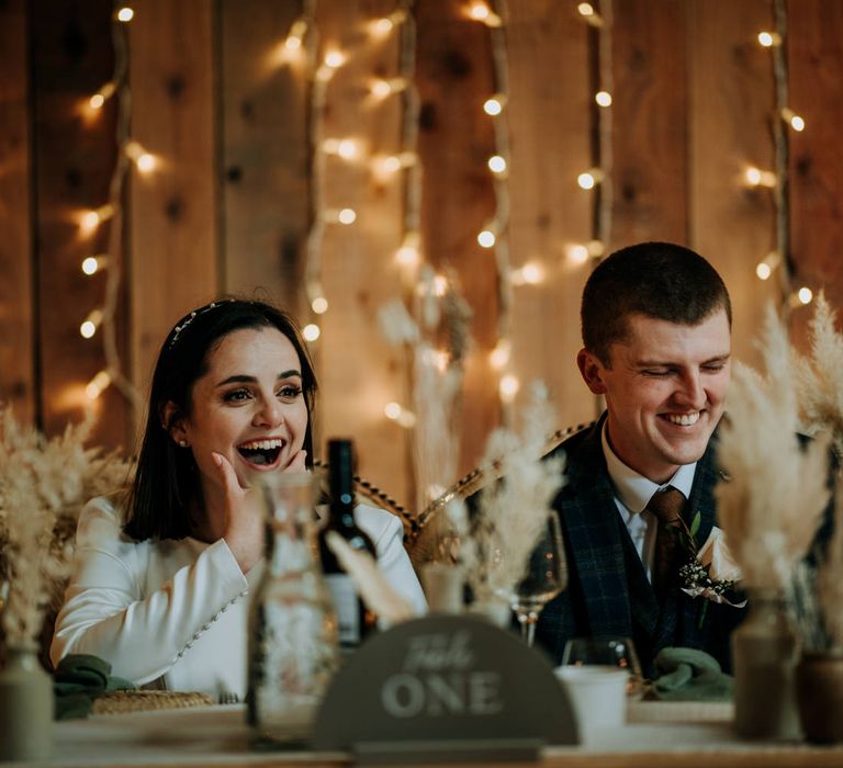 Bride and groom laughing during the speeches at their East Yorkshire Barns wedding reception decorated with fairy lights and pampas grass