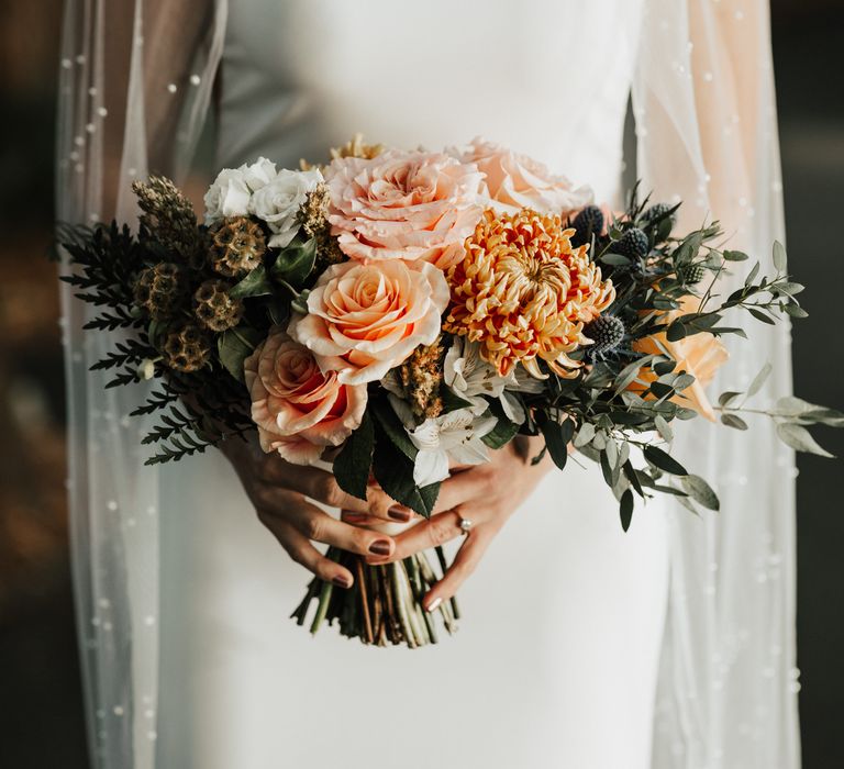 Peach coloured floral bouquet against white bridal gown and pearl veil 