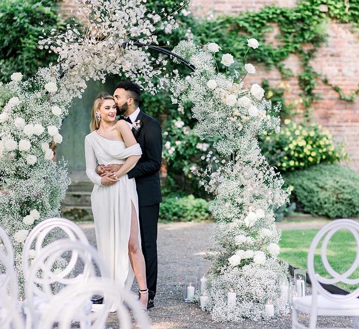 Garthmyl Hall wedding inspiration with groom in a tuxedo kissing his bride in a wrap dress with one shoulder under a white flower arch 