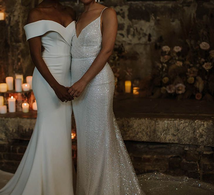 Brides in fitted Made With Love Bridal gowns with off the shoulder detail and sparkly fabric holding hands at their candlelit altar 