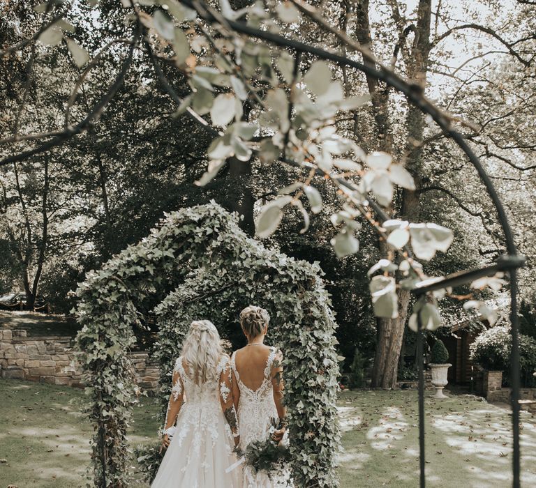 Two brides walk under ivy covered arches at Crab & Lobster fairytale wedding. 