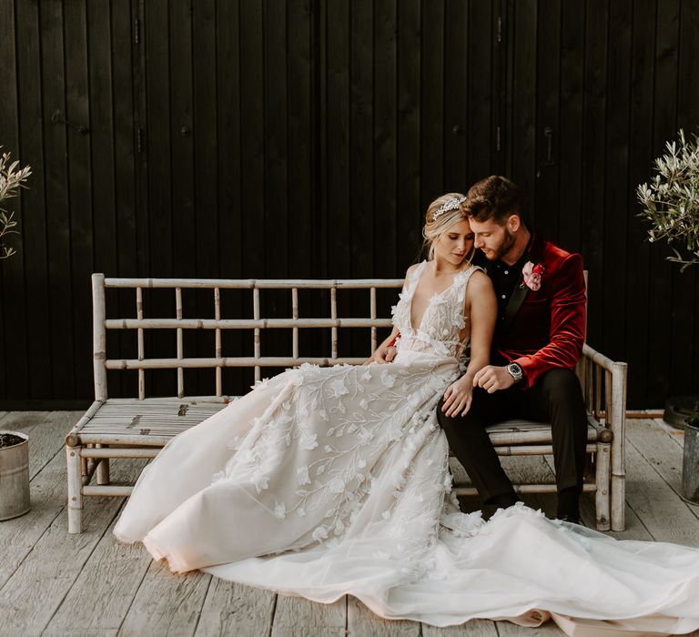 Groom in a red velvet tuxedo jacket embracing his bride on a bench in a lace princess wedding dress