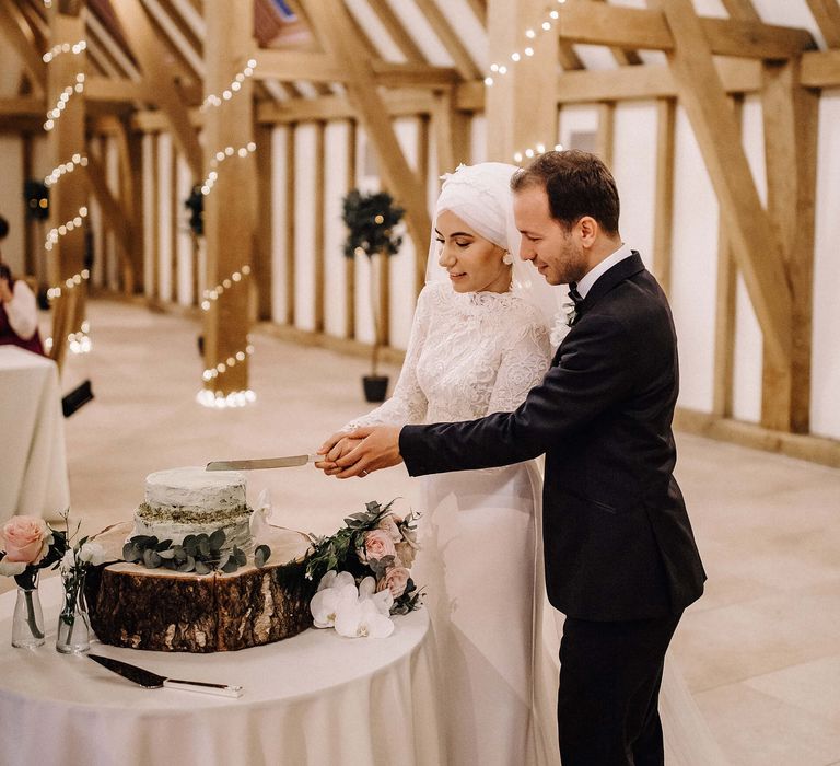 Bride and groom cut the pale green two-tier wedding cake on a tree trunk stand together in Old Kent Barn